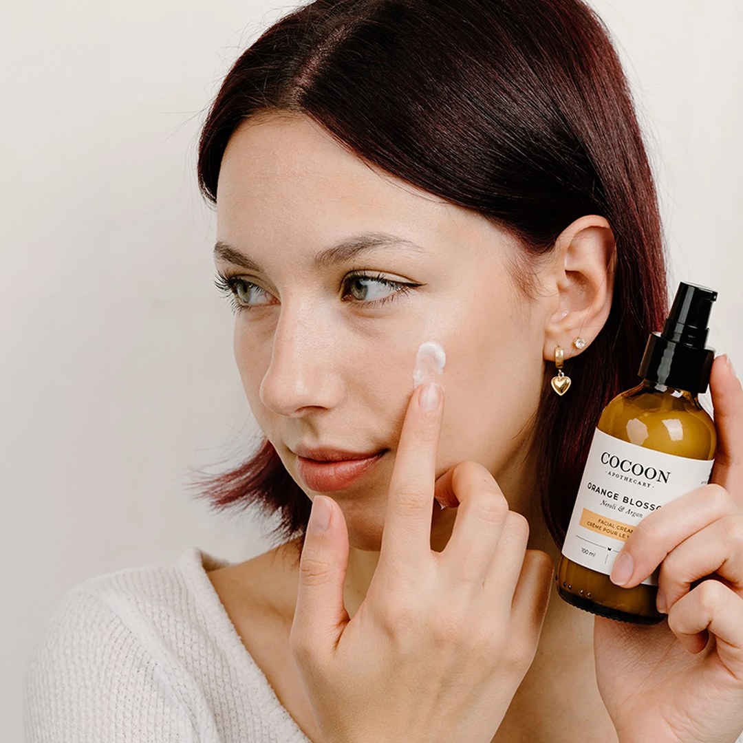 image: a model applying cocoon apothecary's orange blossom facial cream from a bottle.